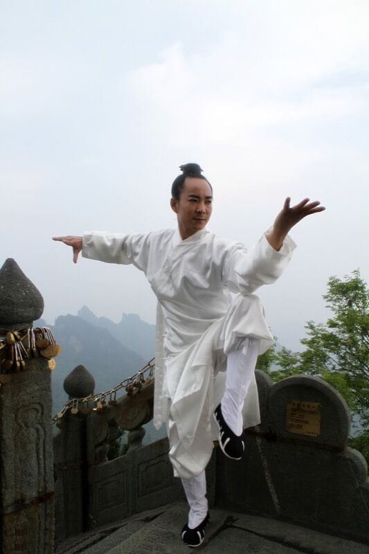 a man in a white traditional chinese martial arts uniform stands on a mountainside with a leg raised. he is practicing a martial art with his hands outstretched. he is wearing black shoes with white soles. the background shows mountains and a cloudy sky.