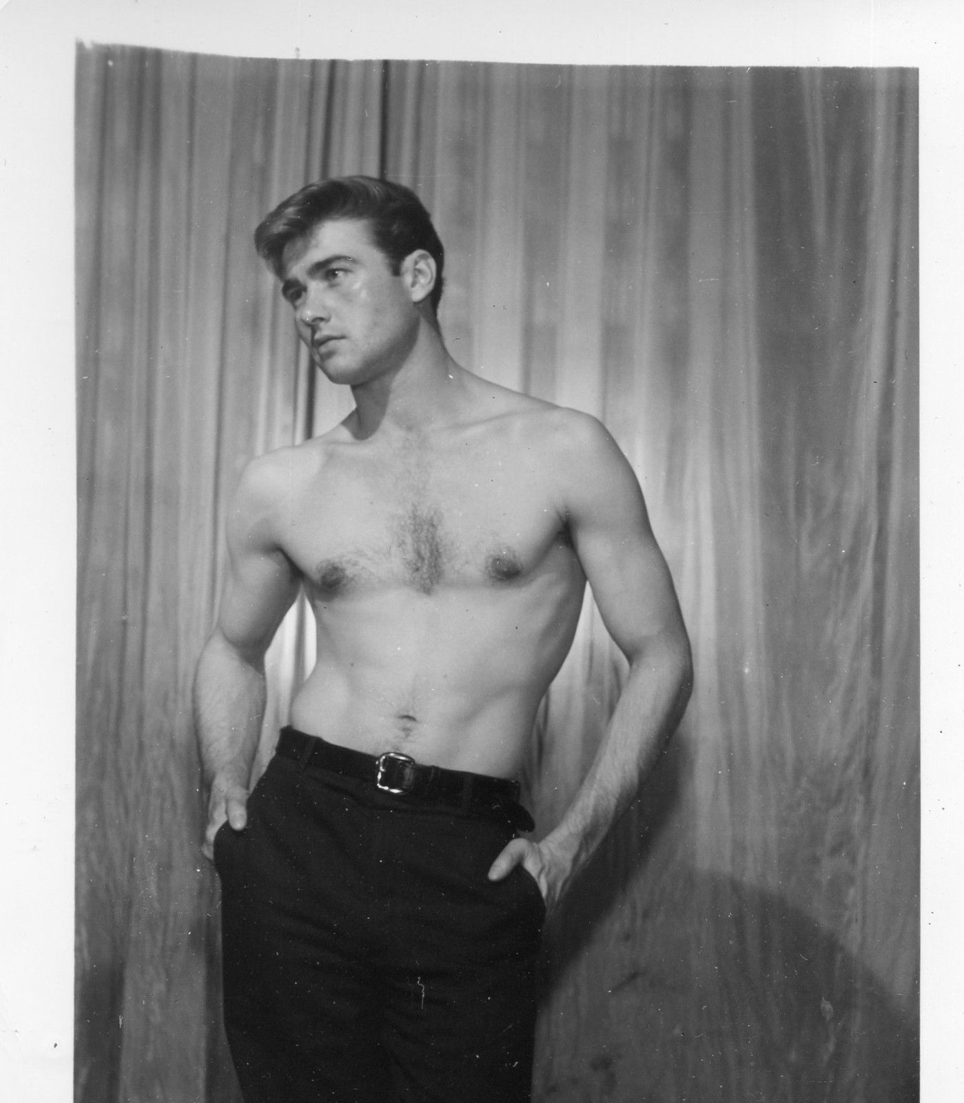he image is a black and white portrait of a muscular young man standing in front of a textured backdrop, likely in a studio setting. he is shirtless, showcasing his physique, and his hands are casually tucked into the pockets of his darkcolored trousers. the man has a neutral expression on his face and his gaze is directed slightly offcamera, creating a sense of contemplation. the lighting is soft and diffused, highlighting the contours of his body and creating a classic, timeless feel.