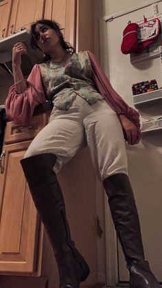 a young woman poses for a mirror selfie in her kitchen. she is wearing a stylish vintageinspired outfit.  she wears a floral patterned vest, a white buttondown shirt,  highwaisted white pants and kneehigh brown boots. she is leaning against the kitchen counter.  the lighting is artificial indoor lighting, creating a warm and inviting atmosphere. the low camera angle adds a sense of drama and power to the image.