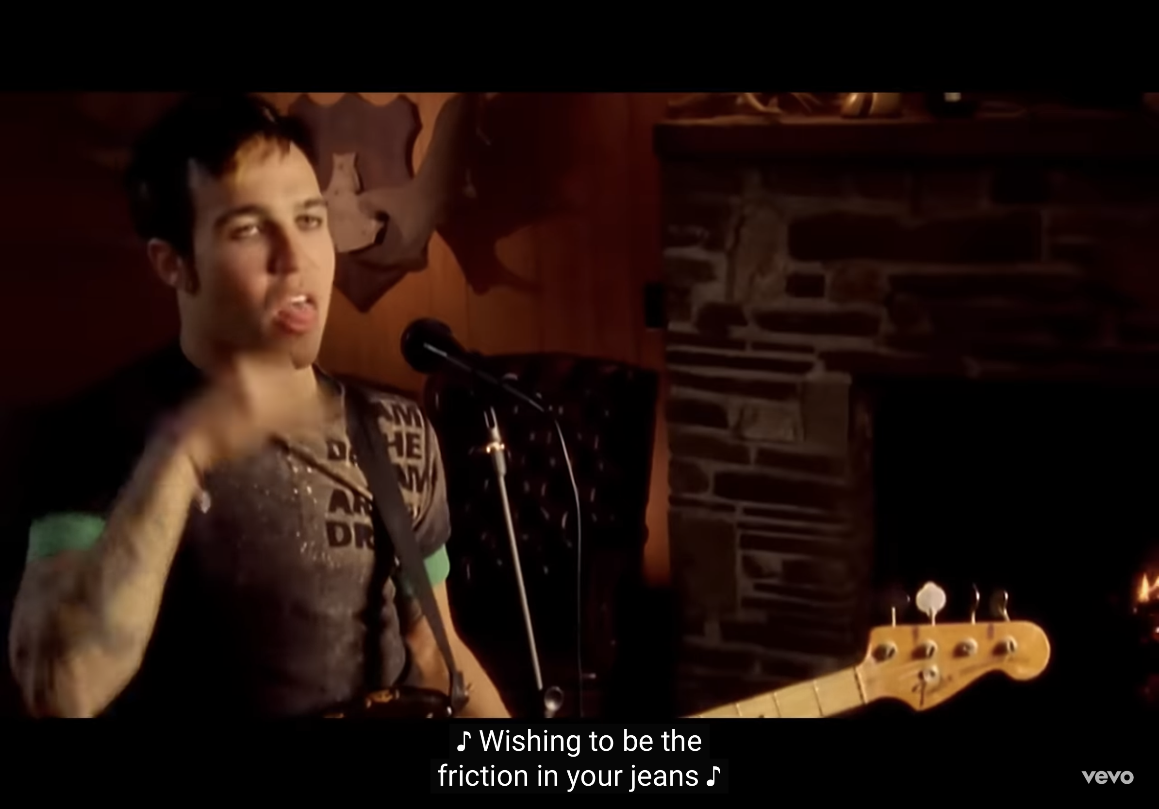 a man with dark hair wearing a grey tshirt and green undershirt is singing and playing a bass guitar. he is standing in a dimly lit room with a stone fireplace behind him.  the lyrics "wishing to be the friction in your jeans" are displayed at the bottom of the image.
