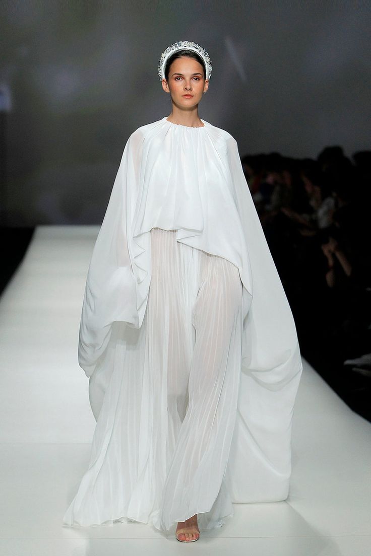 a model in a flowing white dress is walking down a runway in an elegant pose. she is wearing a headband and jewelry, and her long hair is parted in the middle. the dress has a long flowing cape, and the skirt is pleated. the background is blurred and out of focus.