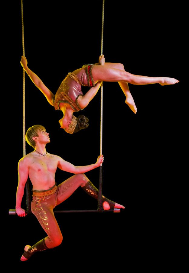 two circus performers are hanging from a trapeze. the female performer is hanging upside down, while the male performer is hanging rightside up. they are both suspended high in the air and are about to perform a complex acrobatic move.