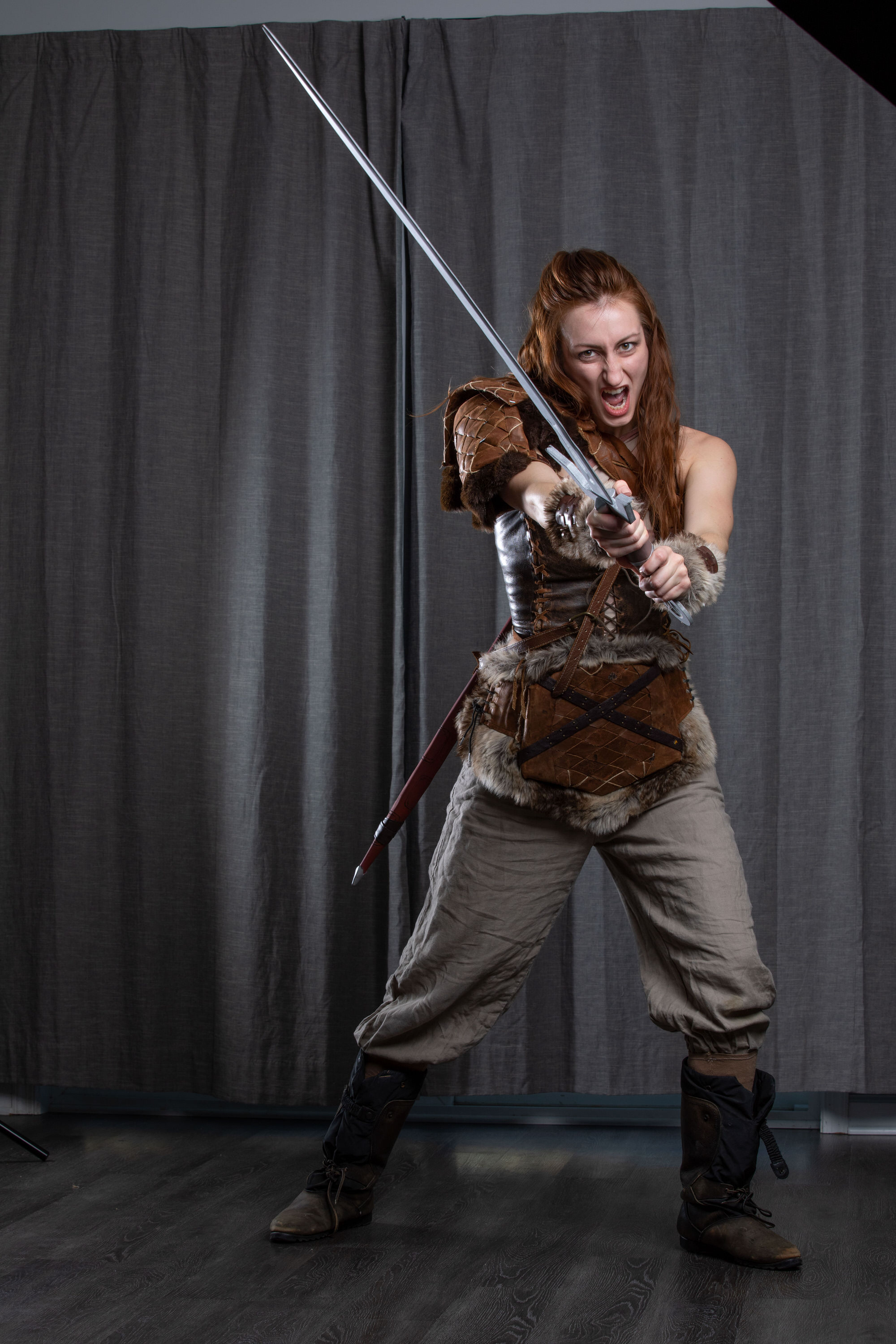 a woman with long red hair pulled back in a halfponytail stands with her legs apart and torso facing the viewer.  she wears a fauxleather and fur jerkin over a longsleeved, light brown tunic and matching pants.  a scabbard hangs on her left hip.  she wears brown boots that rise to midcalf. her mouth is open in a yell, and she grips a sword with both hands, positioned to block an attack coming from the right. the background is a grey curtain, and the dark floor is slightly reflective.