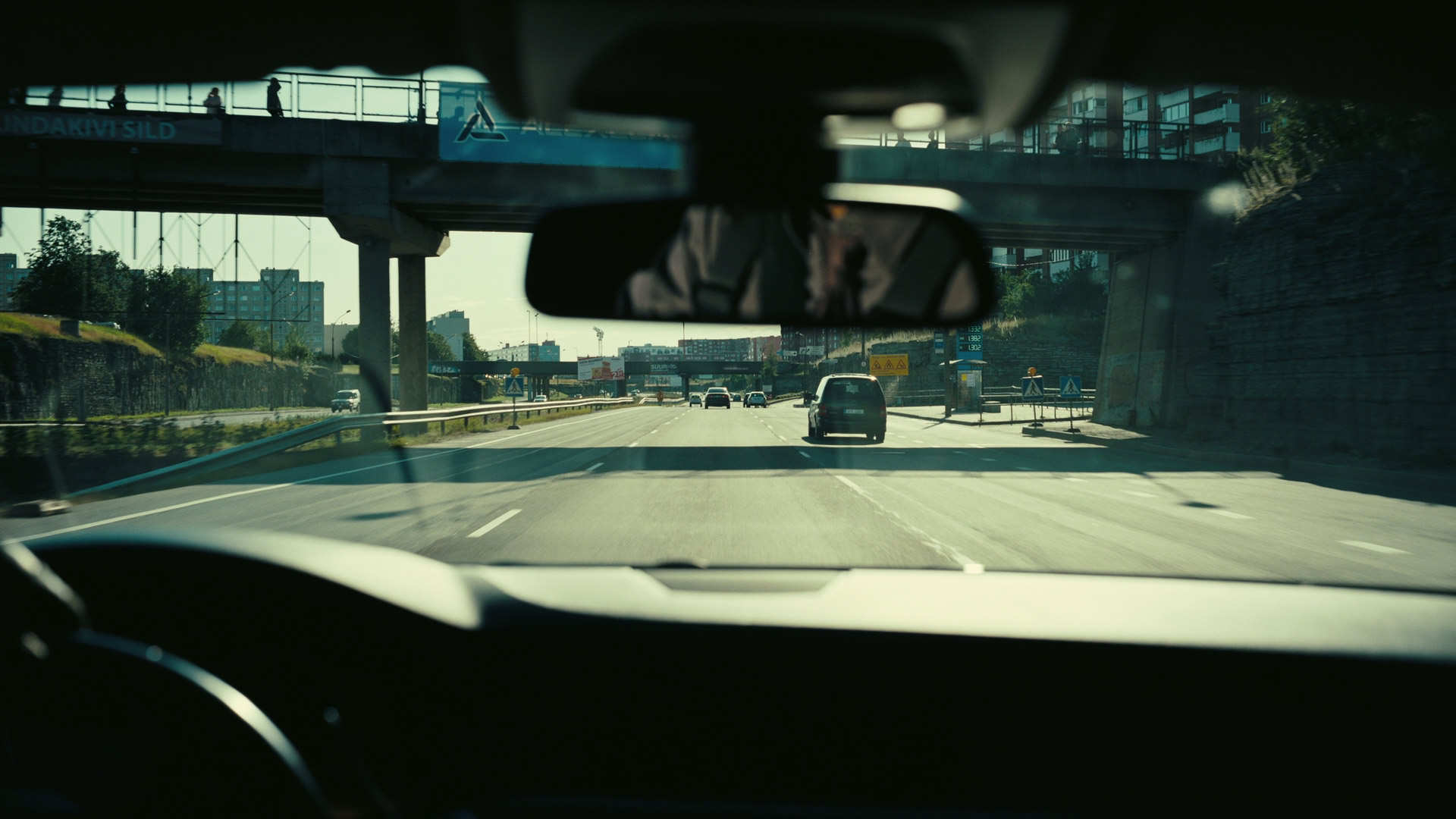 a view from inside a car driving on a highway in the city. the car is traveling under a bridge and there are other cars in the distance. the cityscape is visible in the background, with buildings and trees lining the highway. the sun is shining and the sky is blue. the driver's perspective is shown through the windshield, with the rearview mirror reflecting the cars behind. the steering wheel and dashboard are also partially visible in the foreground. the image captures the everyday experience of driving in a city and the feeling of being on the move.