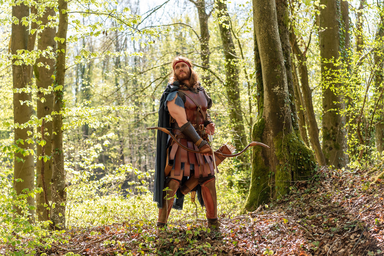 a man dressed in ancient greek or roman warrior attire stands in a sundappled forest.  he is holding a bow and arrow, seemingly ready to shoot. he wears leather armor, a dark cape, and a red headband. the setting is a lush, green forest with tall trees and sunlight filtering through the leaves. the overall image evokes a sense of mythology and historical reenactment.