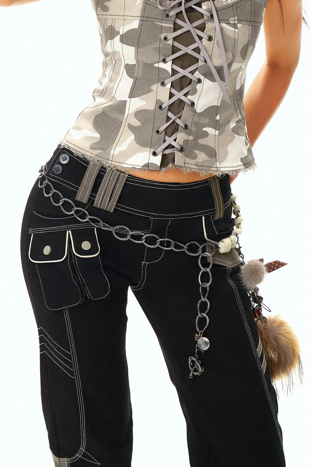 closeup shot of a woman's torso and legs, showcasing her outfit. she's wearing a camouflagepatterned corset top paired with black cargo pants featuring white stitching details.  a long metal chain belt with various charms and pendants is prominently displayed around her waist. the background is pure white, focusing all attention on the clothing and accessories. the image highlights the stylish and trendy nature of the attire.