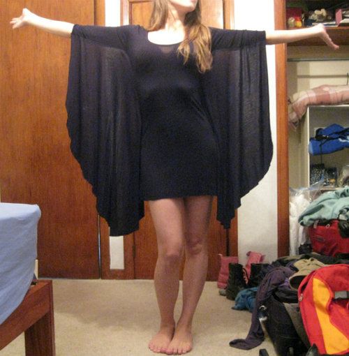 a woman is standing in a bedroom, wearing a black dress with bat sleeves, with her arms outstretched. the background shows a wooden door, a messy wardrobe and clutter on the floor.
