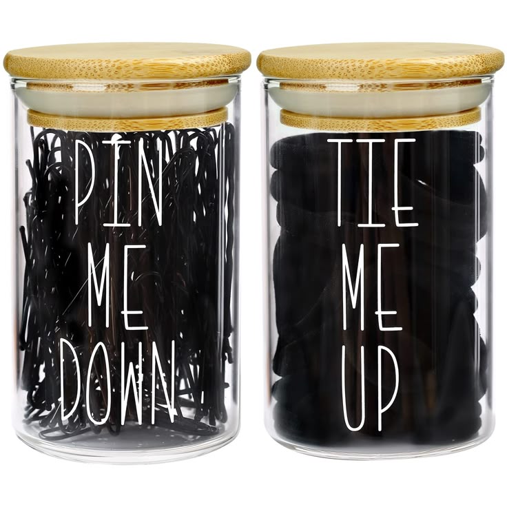 two glass jars with bamboo lids sit sidebyside. the jar on the left is labeled "pin me down" and filled with black bobby pins. the jar on the right is labeled "tie me up" and filled with black hair ties.
