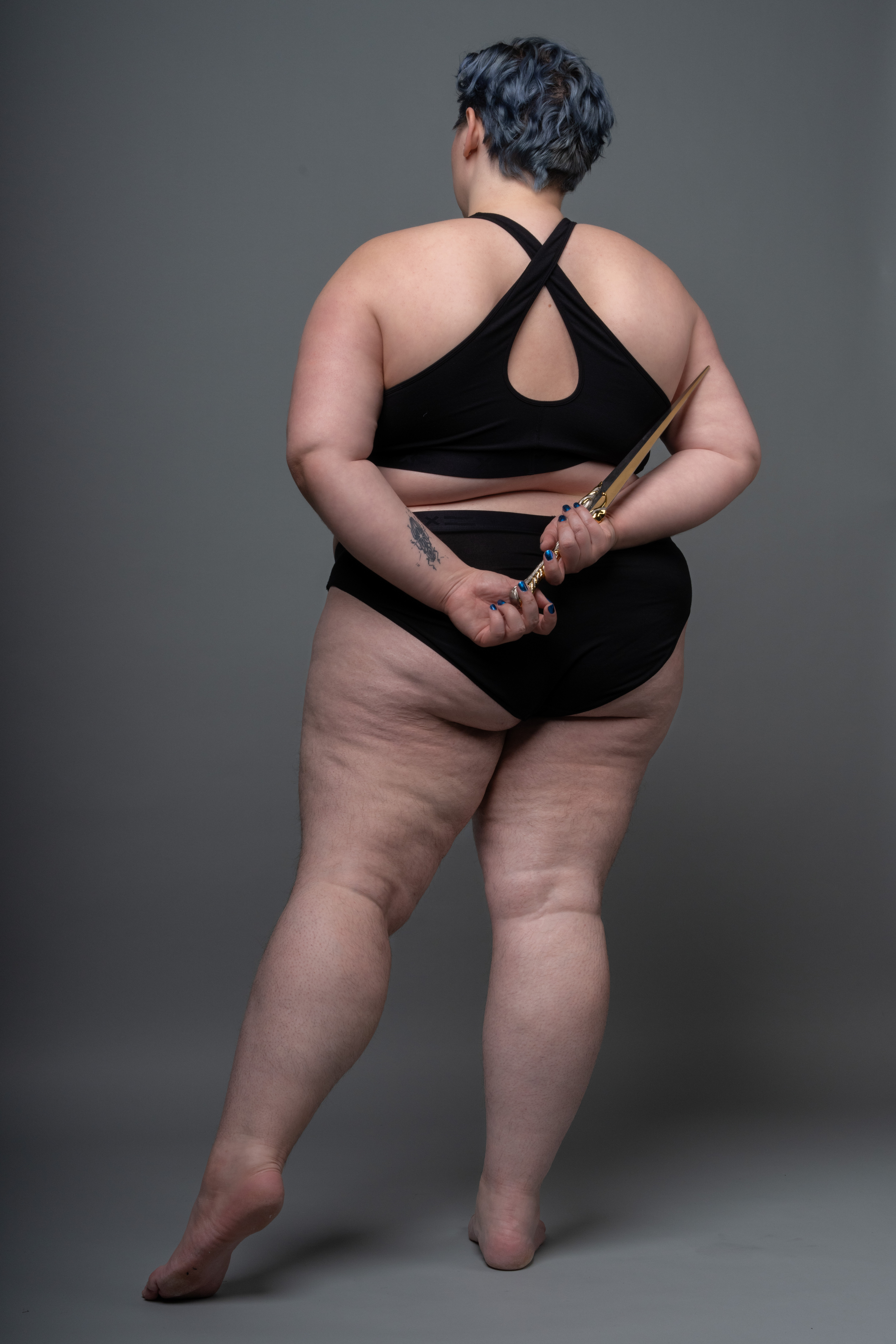 a plussize woman stands with her back to the camera. she is wearing a black bra and black underwear. she has short blue hair and a tattoo on her left arm. she is holding a pair of golden scissors behind her back. the background is a plain grey. she is barefoot.