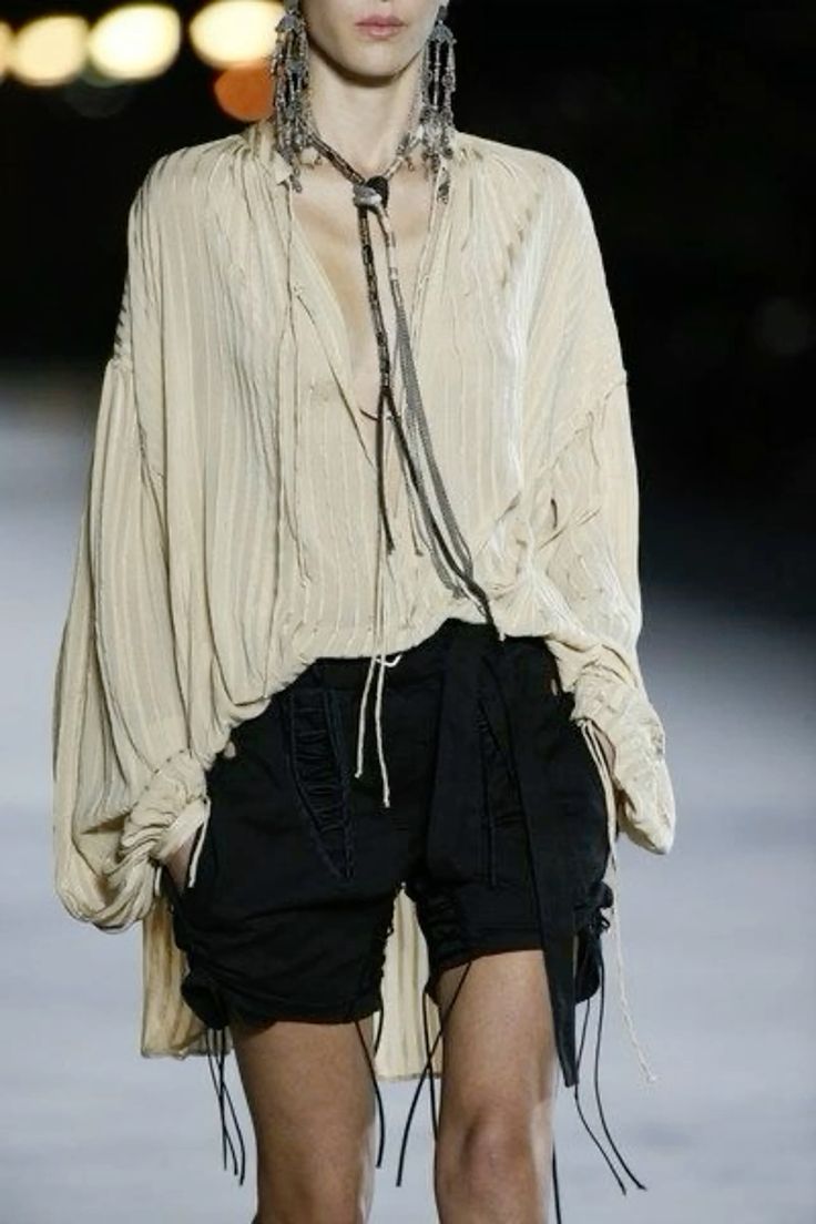 a fashion model walks down a runway in a white, longsleeved, loose shirt with black shorts and several long necklaces. she has long, silver earrings on.