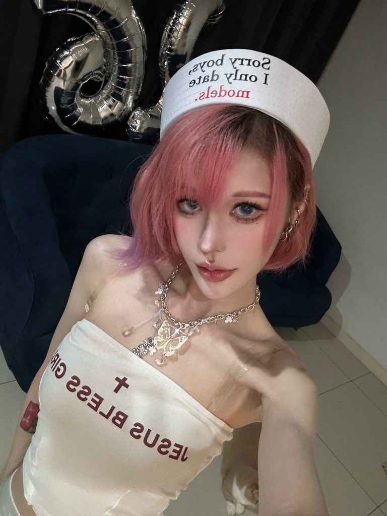 a closeup selfie of a young woman with vibrant pink hair. she's wearing a white sailor hat that reads "i only date models, sorry boys," a white crop top with "jesus bless girl" written on it, and a silver necklace featuring butterfly pendants. the background includes silver balloons, suggesting a birthday celebration.  the overall style is alternative or egirl fashion.  the image is a selfie taken at what looks like a home setting.