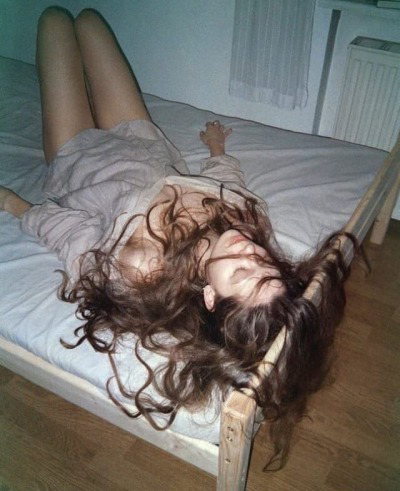 a woman is lying on a bed with her legs up in the air. she has long, brunette hair and is wearing a lightcolored shirt. the bedroom has white walls and a wooden floor.