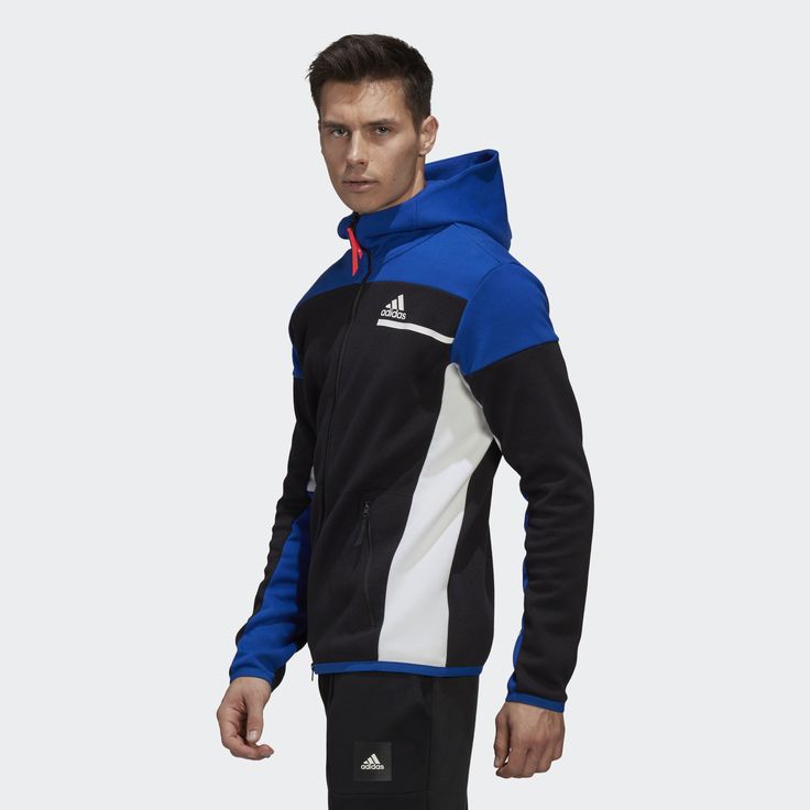 a man wearing a blue, black, and white adidas jacket with a zipper and hood. the jacket features a white adidas logo on the left chest and a black adidas logo on the left hip. the man is standing with his arms at his sides. he is looking at the camera.