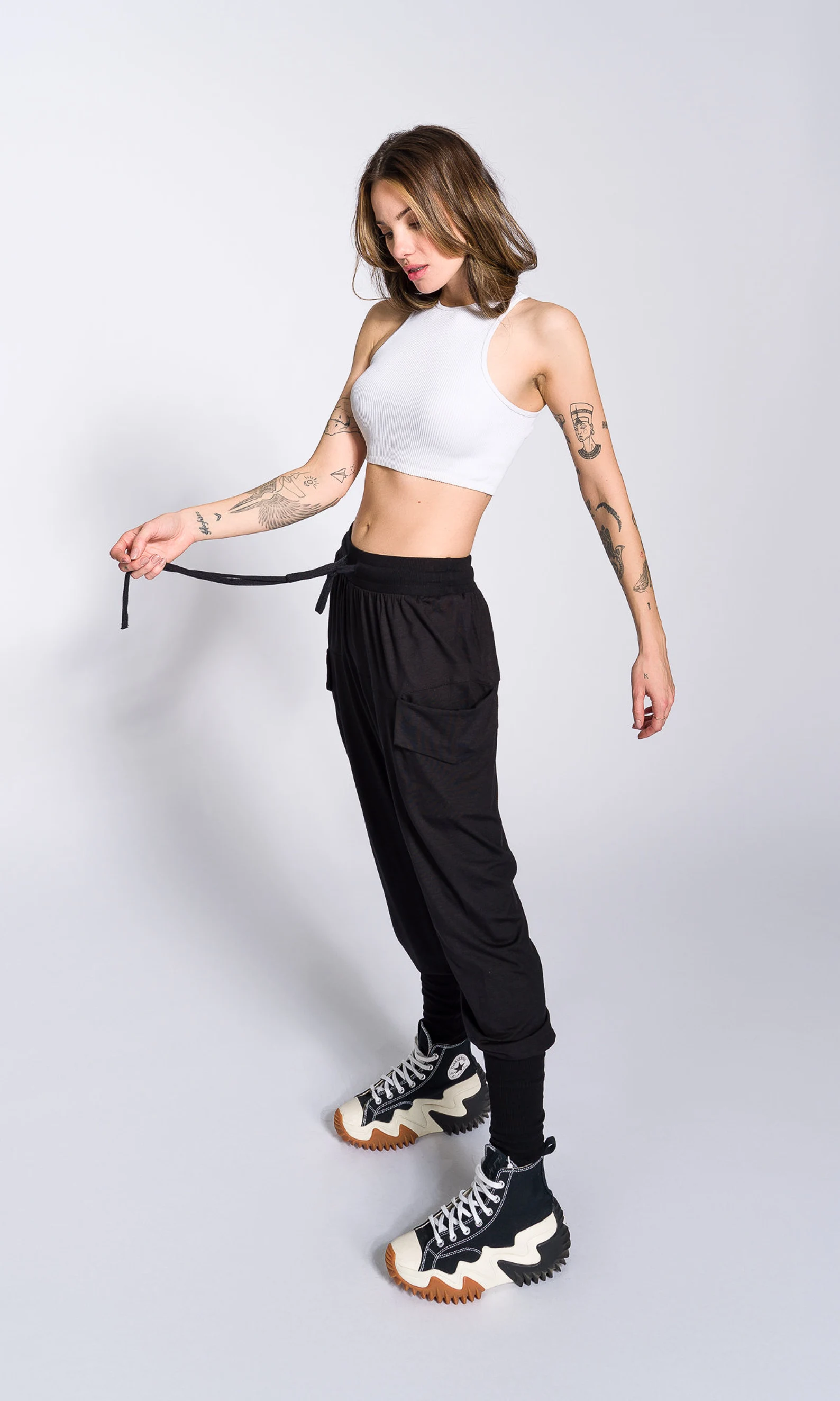 a fullbody shot of a young woman posing in a studio setting. she is wearing a white crop top, black cargo pants, and black and white converse run star hike sneakers. she has several tattoos on her arms. the background is plain white. the overall style is casual and trendy.
