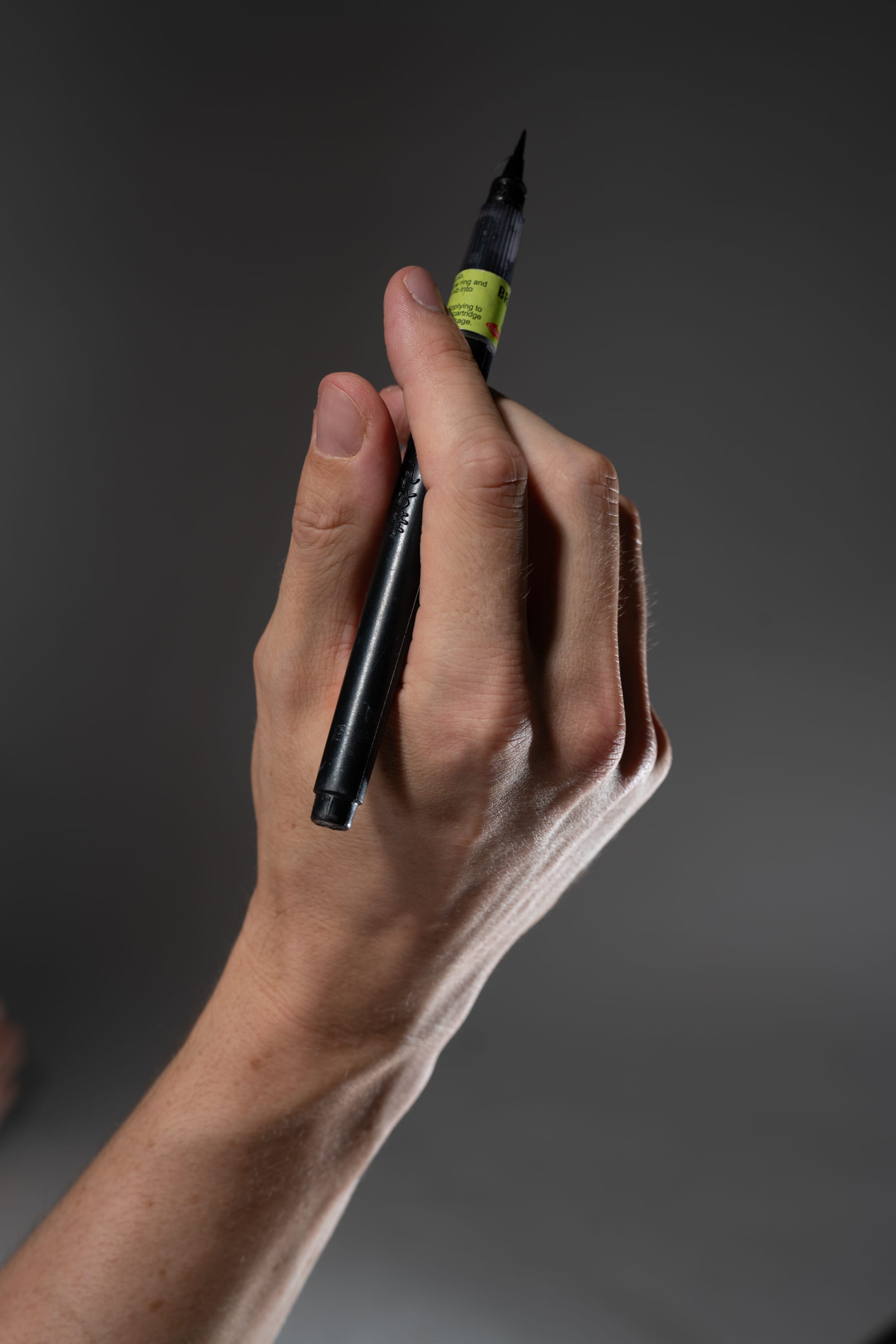 a closeup of a hand holding a black brush pen against a gray background. the pen is held between the thumb and index finger, with the other fingers curled around it. the hand is pale and has visible veins. the lighting is soft and even.
