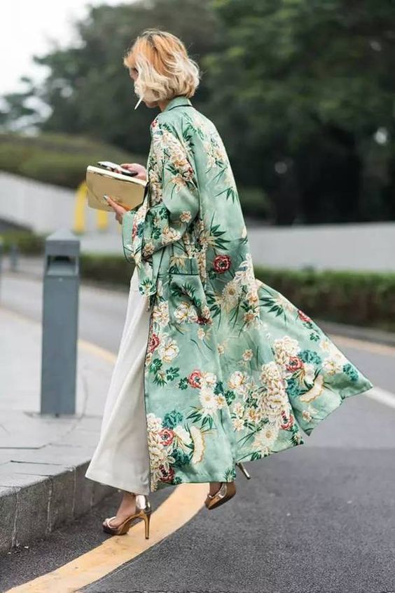 a stylish blonde woman is walking down a city street. she is wearing a long, flowing green kimonostyle coat with a floral print.  underneath, she wears wideleg white pants and gold highheeled shoes. she carries a gold clutch. the overall style is elegant and chic. the setting is an outdoor street scene with a grey pavement and some greenery in the background.  the image is welllit and provides a good view of the woman's outfit and style.