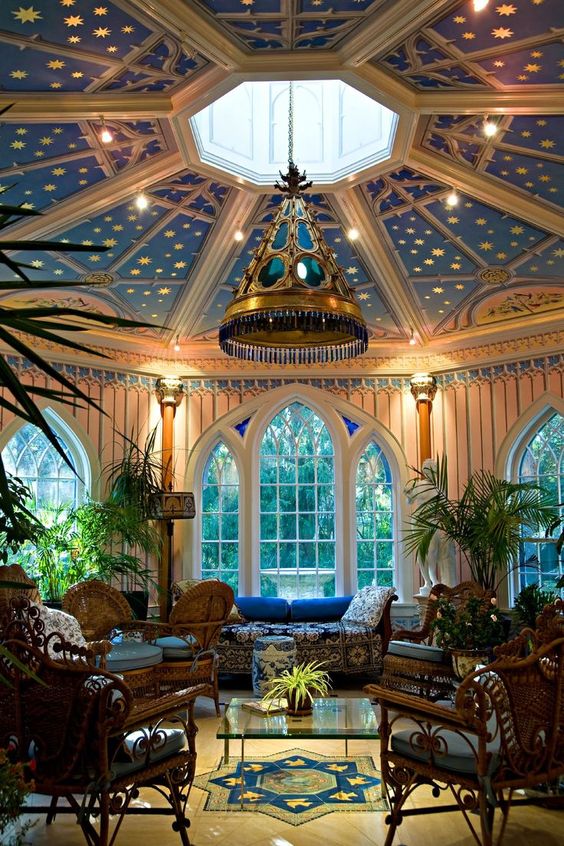a vibrantly colored sunroom featuring a blue ceiling with gold stars, a large skylight, and tall arched windows. the room is furnished with wicker chairs, a blue couch, and various potted plants, creating a tranquil and inviting atmosphere. a large ornate chandelier hangs from the center of the ceiling.