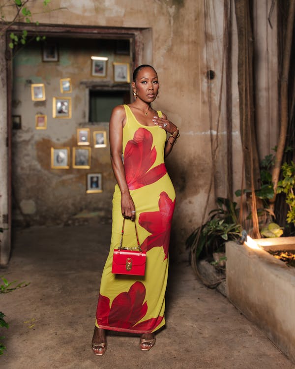 regina king is standing in a doorway of an old building. she's wearing a stunning yellow maxi dress with a red floral print, gold jewelry, and red heels. she carries a small red handbag. the setting is outdoors, with plants and aged stone walls visible. the image is a sophisticated and elegant portrait.