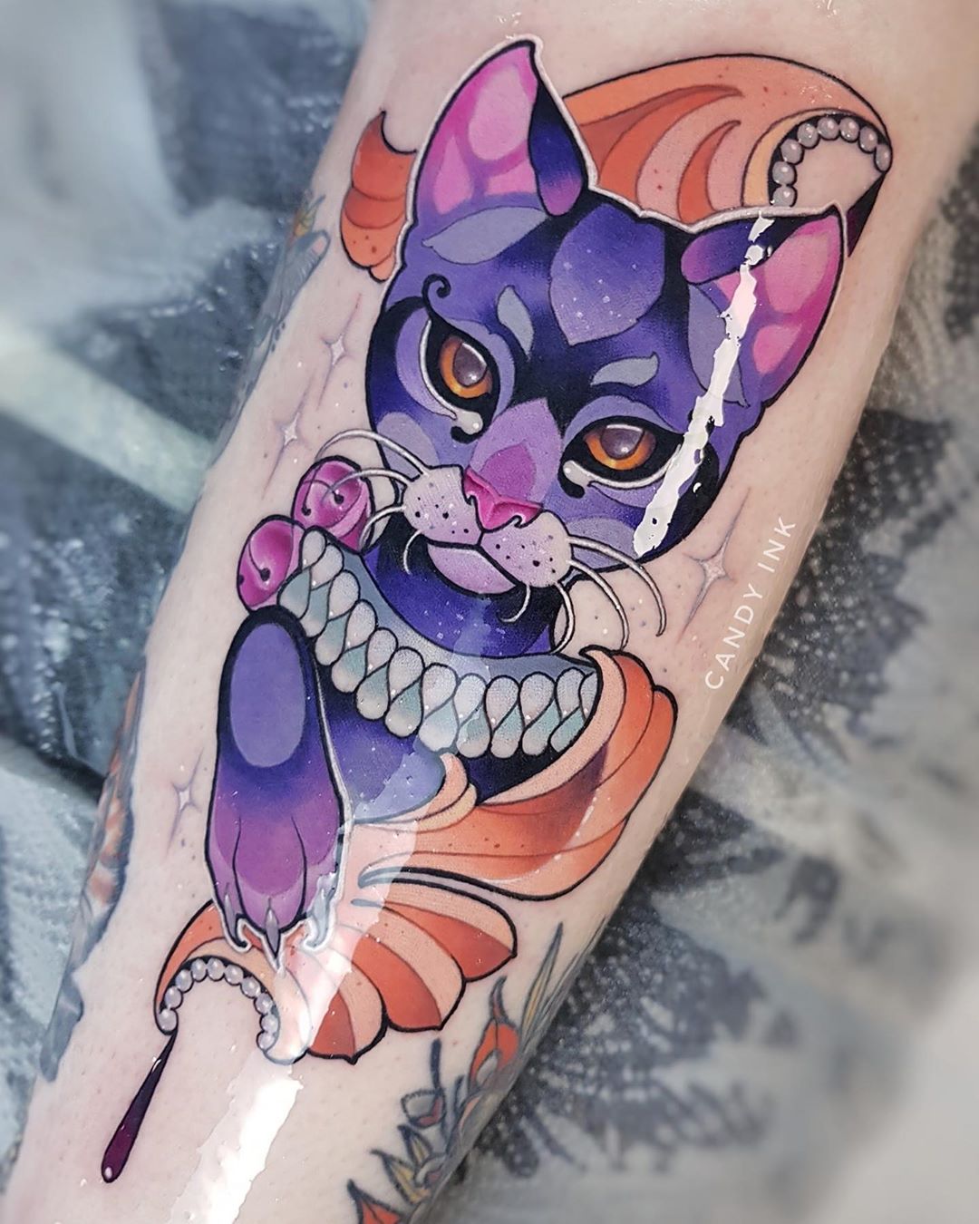 a tattoo of a regallooking purple cat with orange and pink accents. the cat has a pearl necklace and there are other jewels in the design. the tattoo is on someone's inner forearm, and you can see the tattoo artist's station in the background. the tattoo artist's signature, "candy ink" is written on the tattoo.