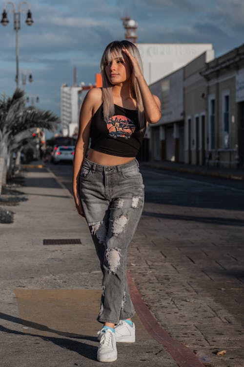 a young woman with blonde hair and bangs is casually posing on a city street. she is wearing a black graphic crop top, light gray ripped jeans, and white sneakers.  her style is relaxed and trendy, showing off a contemporary street style look. the setting is an urban environment, with buildings and palm trees visible in the background. the lighting suggests it's daytime.