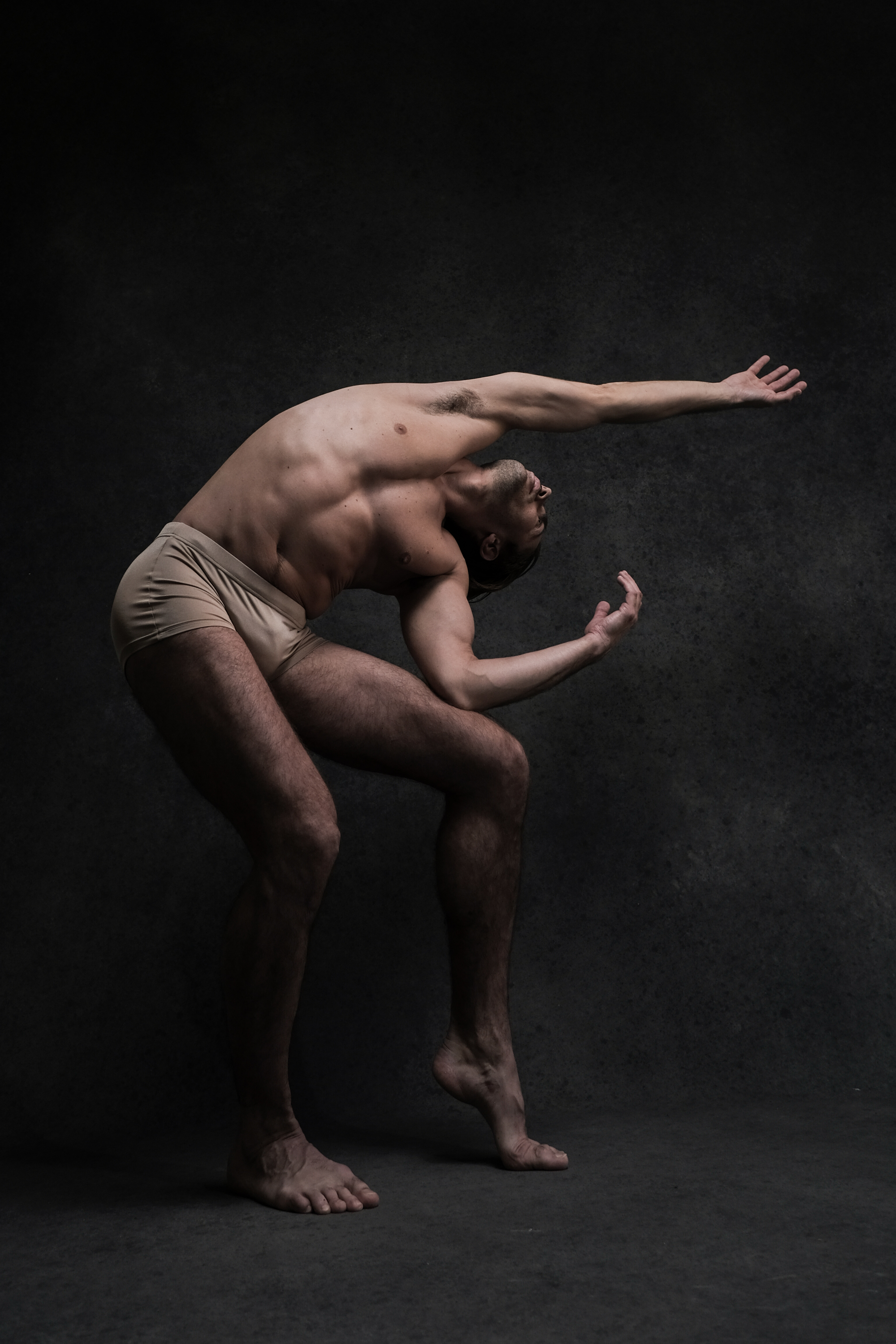 a male dancer in beige underwear is posed dramatically against a dark grey background.  he is leaning backward, his right leg bent, his left leg straight, with his arms stretched out.  the lighting is dramatic, highlighting his muscles and creating shadows on the background.  his facial features are obscured as his head is tilted back.