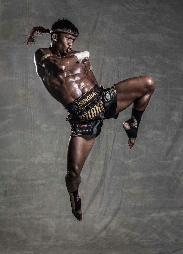 a male muay thai fighter midkick in a grey studio. he is wearing black shorts and a black headband, and he is focused and determined. his muscles are tense and his face is contorted in effort.