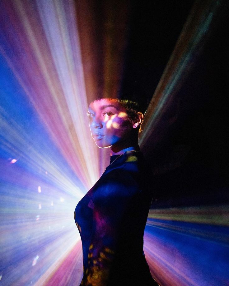 a woman in a dark blue shirt stands in a dark room illuminated by a colorful neon light. her face is half obscured by the light, and she is looking to the left of the frame. the light is emanating from behind her, making her a silhouette. the room is dark and mysterious, and the woman's silhouette is the only thing that stands out. the image is dramatic and artistic, and it creates a sense of mystery and intrigue. the colors in the image are bright and vibrant, and they add to the overall sense of energy and excitement. the woman is standing in a pose that is both confident and mysterious, and she is clearly the subject of the image.  the image could be interpreted as a portrait of a woman, or it could be seen as a more abstract representation of light and color. the image is open to interpretation, and it is sure to spark conversation and discussion.