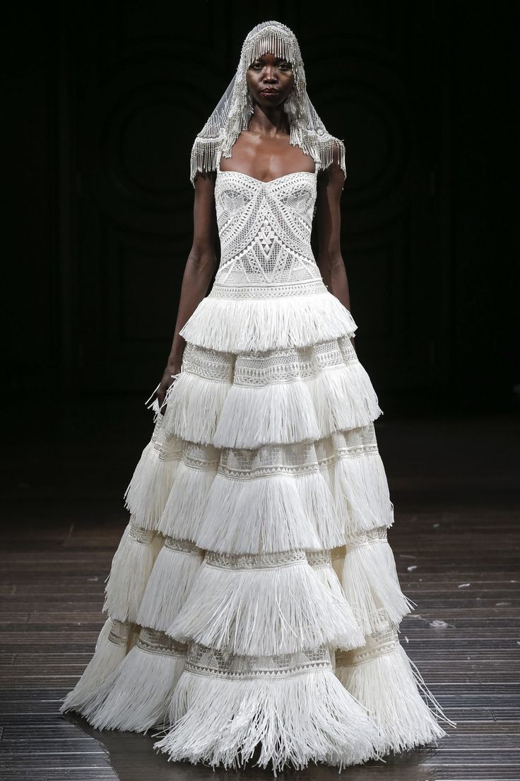 a model walks the runway in a white wedding dress with a lace bodice and a multitiered skirt featuring fringed trim. she is wearing a beaded headpiece and a veil.