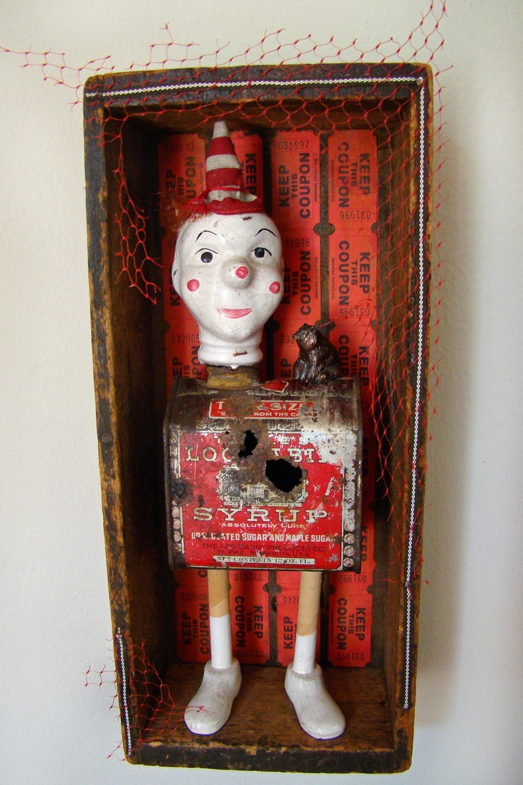 an assemblage art piece inside a rustic wooden shadow box features a whimsical clown figure. the clown's head, with painted features and a red and white striped hat, is perched on a rusty "loc syrup" tin can, which acts as the torso. the can has rusted holes and faded paint. beneath the can are wooden legs with painted socks and shoes. behind the figure are rows of vintage red coupons. a small dark figure, possibly an animal, sits on top of the syrup can. the shadow box is lined with red netting, adding a textural element to the piece. the overall effect is a blend of folk art and recycled materials, creating a quirky and imaginative art doll.