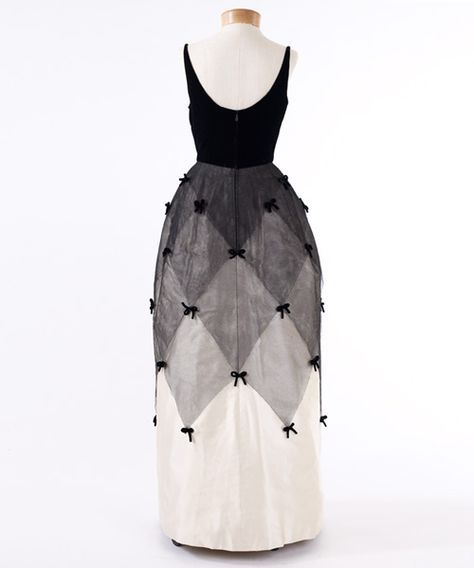 a formal gown on a mannequin, featuring a black velvet sleeveless bodice and a long skirt. the skirt has a diamond pattern created by a sheer fabric overlay with small black bows attached at the corners of each diamond. the lower part of the skirt is white. the mannequin is facing away from the viewer.