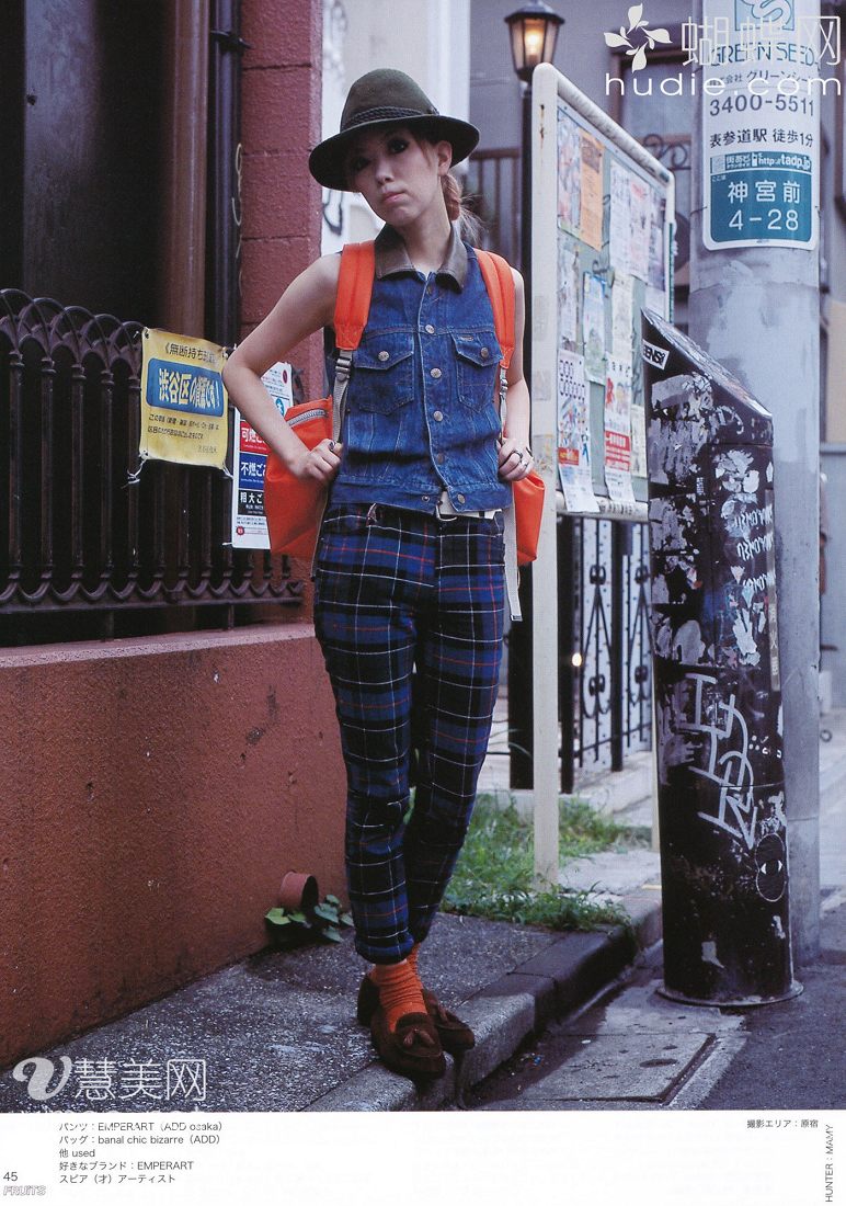 a young asian woman is posing on a city street, showcasing a stylish outfit. she wears a denim vest over a white shirt, paired with blue plaid pants. her accessories include a dark green hat, an orange backpack, and brown shoes with orange socks. the photo appears to be taken for a fashion magazine or website, as suggested by the text overlay featuring brand names and contact information. the overall ambiance is casual and trendy, reflecting a contemporary urban street style.