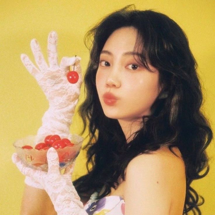 a woman with dark, wavy hair is posing against a yellow background. she is wearing lace gloves and holding a bowl of cherries in one hand, while holding a single cherry with her other hand in an "okay" sign. she has a slight pout on her lips. she's wearing a dress, which has a floral pattern.