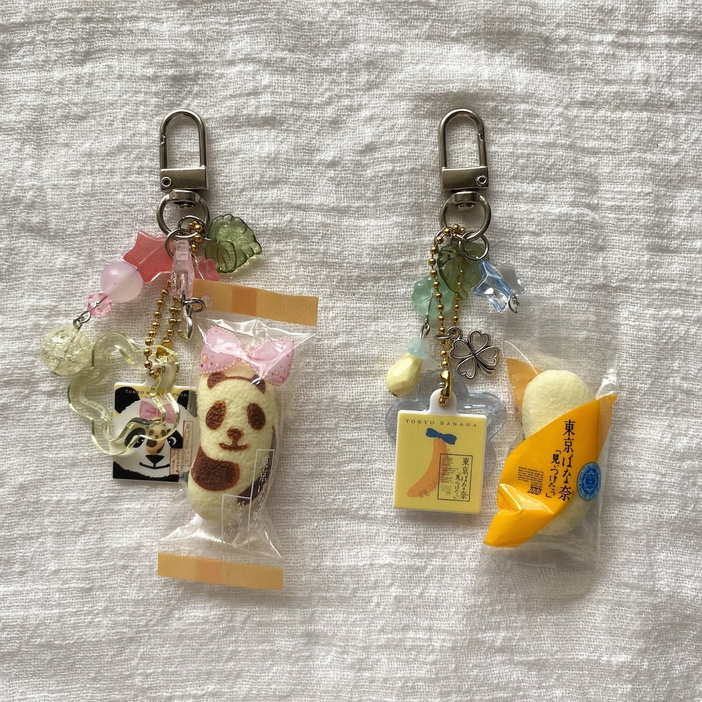 two keychains are displayed on a white textured surface. one keychain features a felted panda treat sealed in a miniature plastic bag, adorned with a pink bow. it is connected to the keychain with beads and leaf charms. next to it, there's another keychain featuring a miniature tokyo banana snack, also in a sealed bag, along with a clover charm, leaf charms, and a tokyo banana logo pendant.