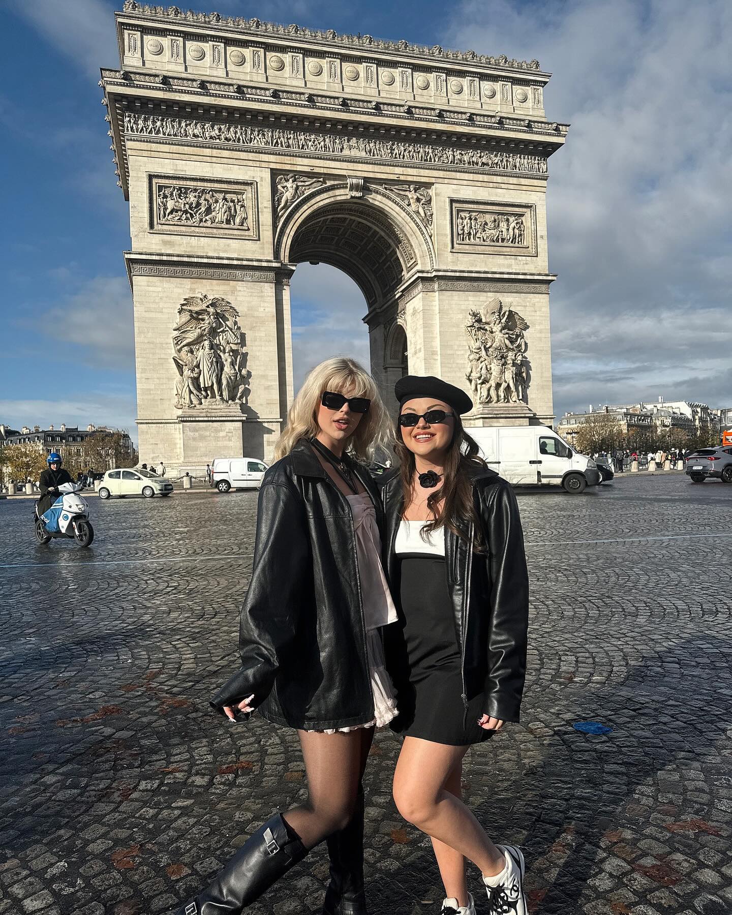 two women stand in front of the arc de triomphe in paris. the woman on the left has blonde hair and is wearing sunglasses, a black jacket, and boots. the woman on the right is wearing a black beret, sunglasses, a black jacket, and sneakers. the arc de triomphe is a large monument in the background. there are other people, cars, and buildings in the background as well. the sky is overcast.