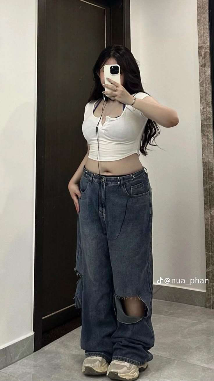 a woman is taking a mirror selfie. she is wearing a white crop top and ripped jeans. she is standing and posing for the photo.