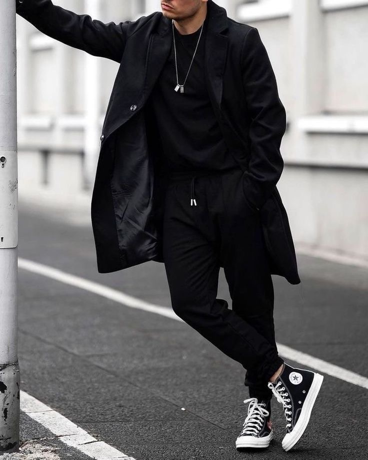 a man wearing a black coat, black trousers and black converse shoes, stands on a paved street. he is looking to the left of the frame. the overall look is trendy and stylish.