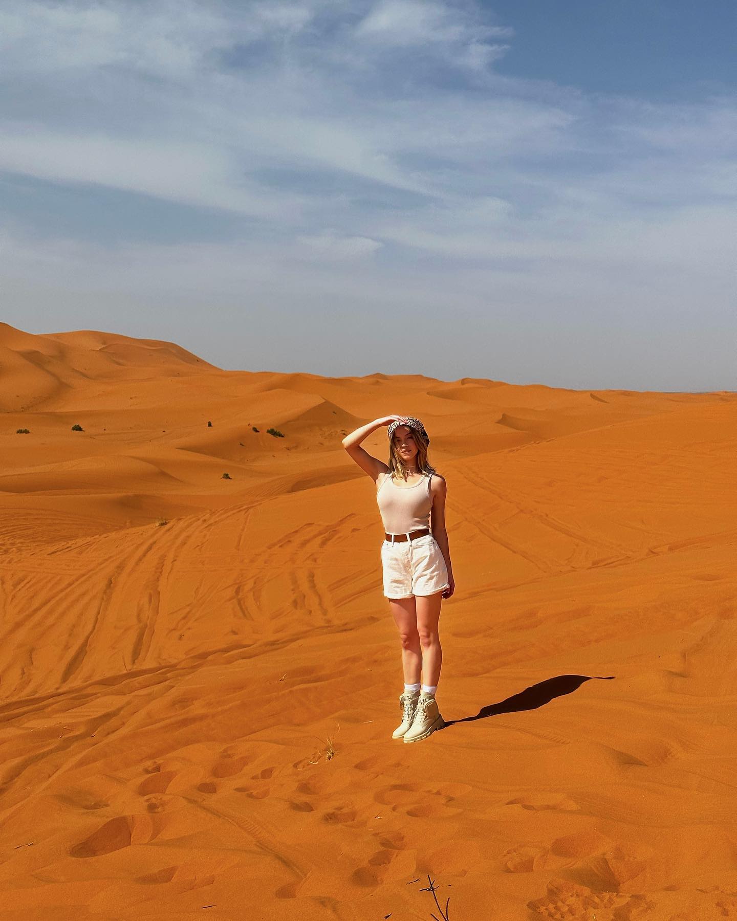 the image shows a young woman standing in a vast expanse of orange sand dunes, characteristic of a desert landscape like the sahara desert. she is wearing white shorts and a white tank top, and beigecolored boots. she has a hat on and one hand is raised to her forehead as if shielding her eyes from the sun. the dunes stretch out to the horizon under a bright, mostly clear blue sky with some wispy clouds. the overall impression is one of adventure and exploration in a dramatic and remote natural setting.  the woman's shadow is cast on the sand beside her.