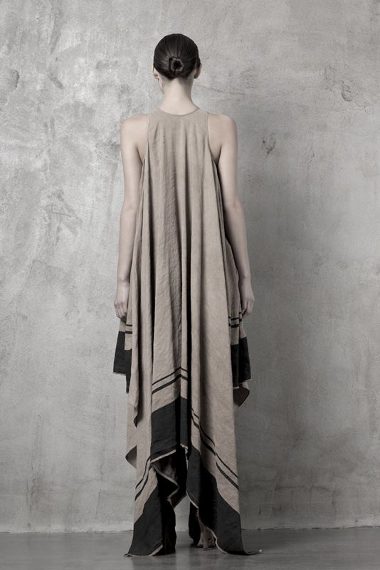 a woman is seen from the back, wearing a long, sleeveless tunicstyle dress. the dress is primarily beige, with bold black horizontal stripes near the bottom, giving it a minimalist and fashionable look. the fabric is draped and loosefitting, possibly linen, creating a sense of flowing movement. the woman's hair is neatly styled in a bun. the background is a simple gray wall, suggesting a studio setting.