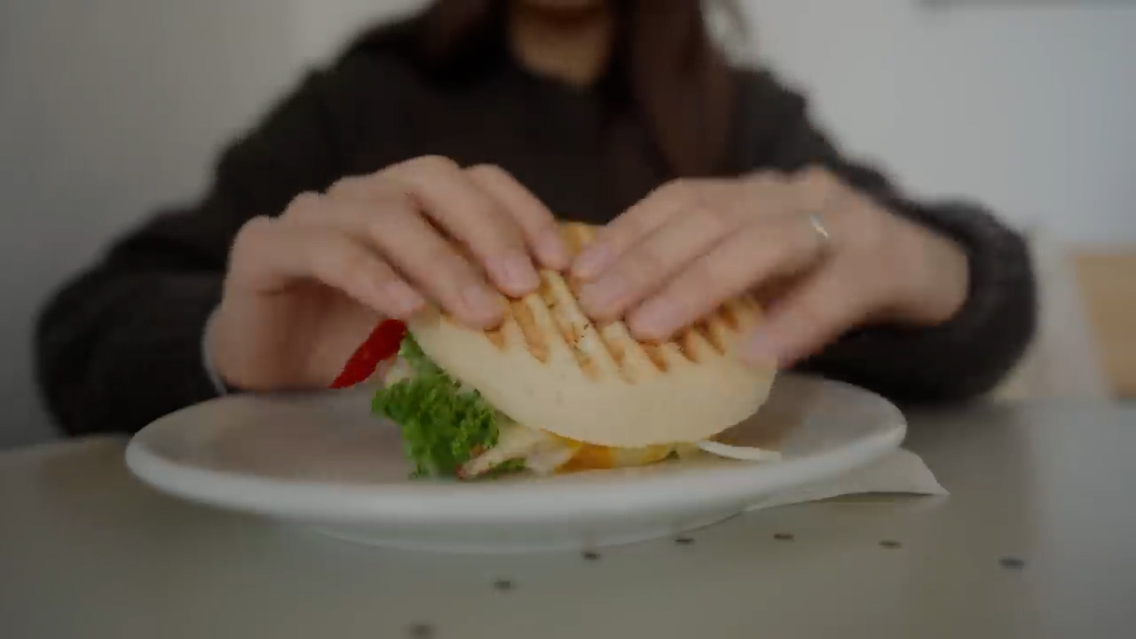 a closeup shot of a person holding a grilled chicken burger with both hands over a white plate. the burger consists of a toasted bun, grilled chicken, lettuce, tomatoes, and sauce. the background is a blurry white wall.