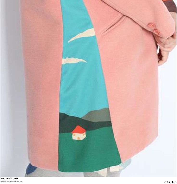 a closeup shot features a pink coat with a unique landscape design panel. the design includes a turquoise sky with white clouds, a green field, and a small house with a red roof. mountains are also depicted. the coat has buttons and a pocket visible, and the seam is a noticeable feature of the design.