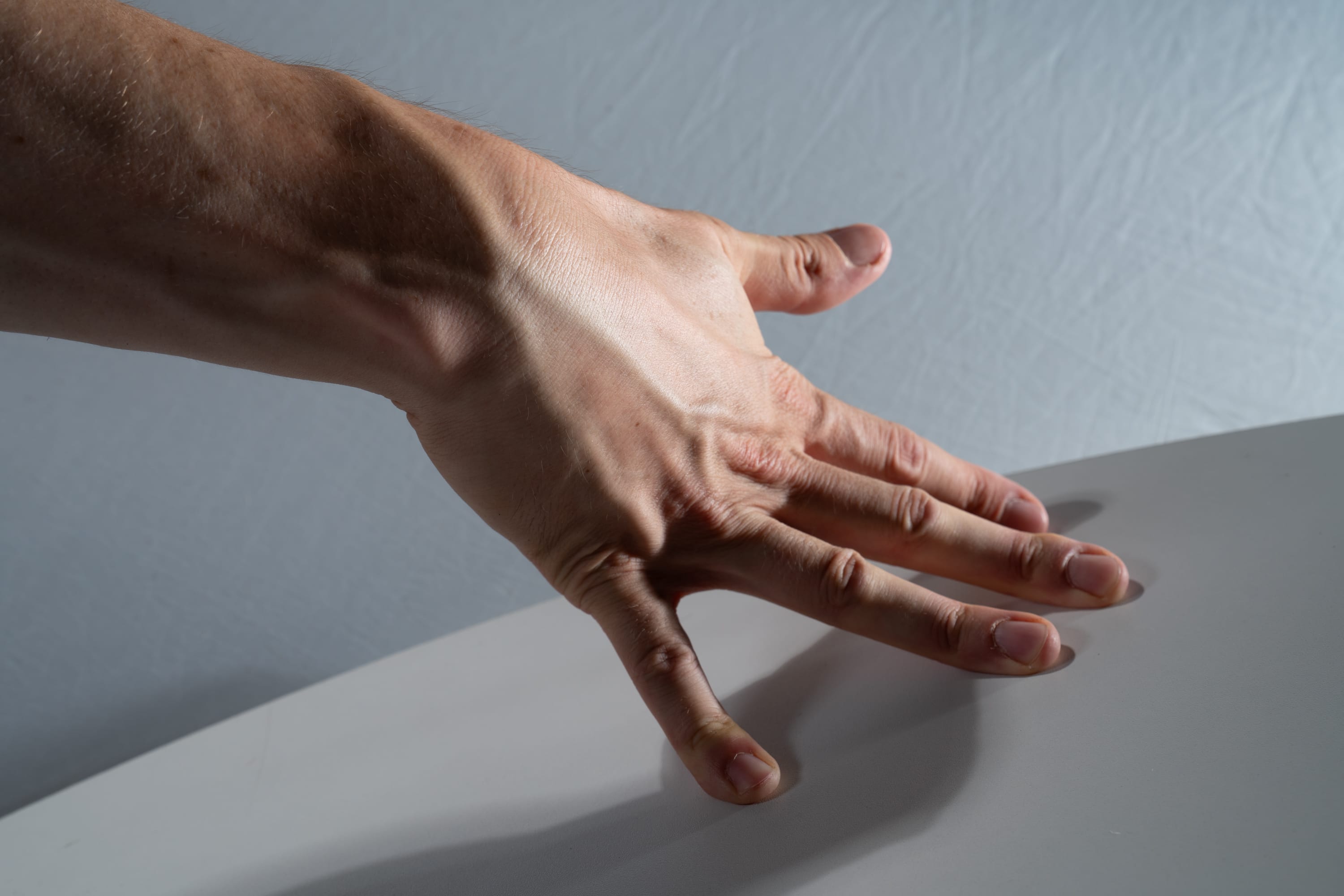 the image shows a closeup of a man's hand resting on a white table. the hand is illuminated by studio lighting, which highlights the veins and tendons in the arm. the background is a plain gray wall.