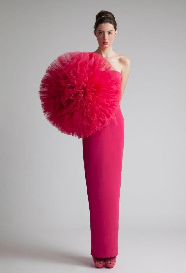 a woman in a pink dress with a large tulle ball on one shoulder, poses against a grey background.