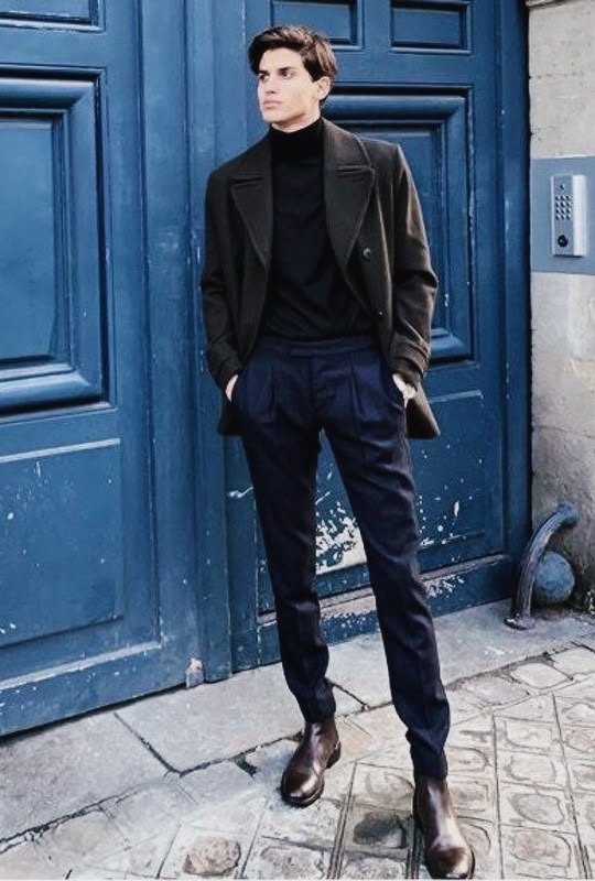 a fullbody shot of a stylish man standing outdoors in front of a blue door. he is wearing a dark green or brown peacoat, a black turtleneck sweater, dark blue pleated pants, and brown chelsea boots. the background includes a textured blue door and a cobblestonepaved street. the man has his hands in his pockets. an intercom system is visible on the wall.