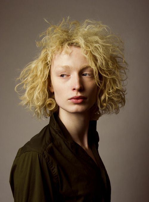 a portrait of a fairskinned model with blonde, curly hair wearing a green shirt, looking away from the camera.