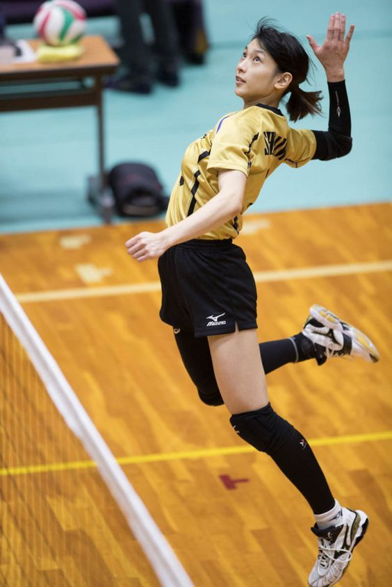 a female volleyball player in midair, about to spike the ball. she is wearing a black and gold uniform and black knee pads. the image is taken from a low angle, emphasizing the height of her jump. the background is a blurred gymnasium with a wooden floor.