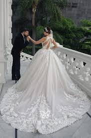 a stunning bride in a longsleeved, white lace ballgown with a low back stands on a balcony with her groom. the groom in a black tuxedo is slightly bent over and looking at the bride while holding her hand.  the bride's dress has a full skirt with delicate lace appliqués and a long train. the setting is outdoors, with lush green plants visible in the background. the overall mood is romantic and celebratory.