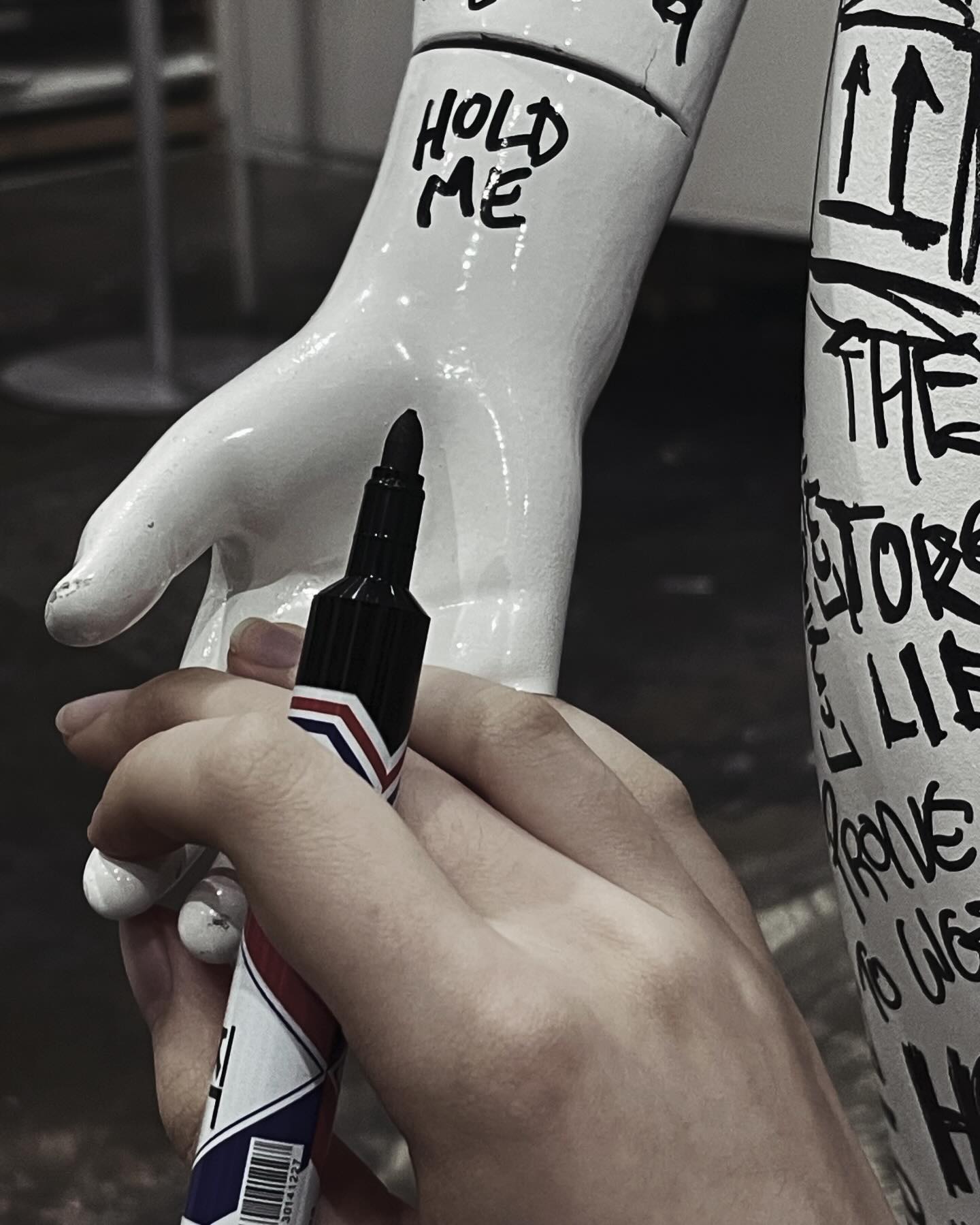 a hand is holding a black marker and writing "hold me" on a white hand.  the hand is covered in graffiti. the hand is in focus while the marker is slightly out of focus.