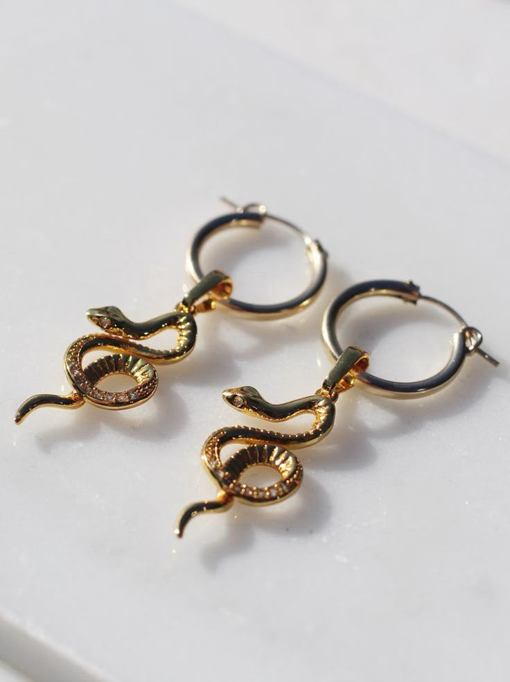 a closeup shot of two gold hoop earrings with snake pendants on a white surface.