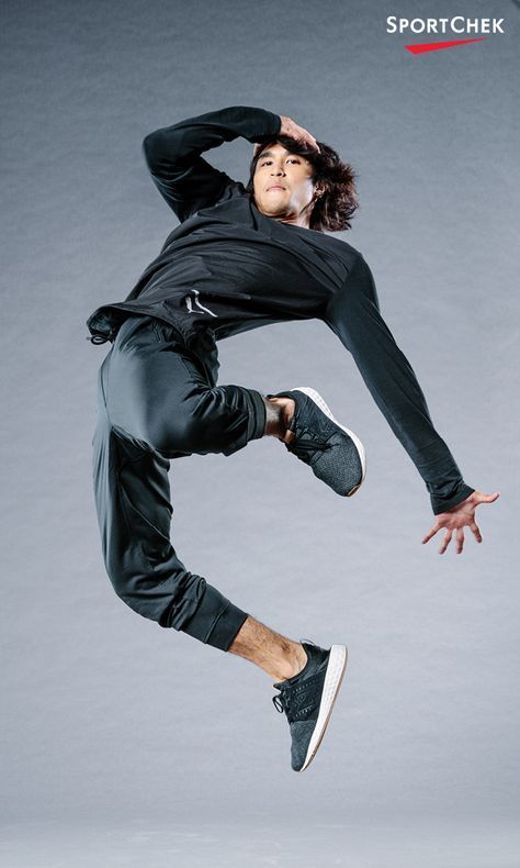 the image shows a young asian man, likely a breakdancer, in midair during a dynamic jump. he is wearing dark grey athletic clothing—longsleeve shirt and sweatpants—and darkcolored athletic shoes. his pose suggests breakdancing or a similar form of hiphop dance. the background is a plain light grey.  the sportchek logo is visible in the upper right corner. the overall impression is one of athleticism, energy, and movement.