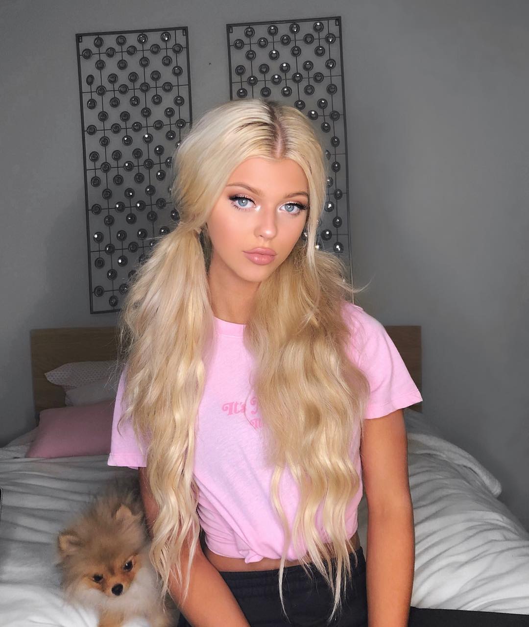 a young woman with blonde hair styled in pigtails poses in a pink tshirt and black shorts in a bedroom setting. a small pomeranian dog is on the bed beside her. wall art hangs behind her bed.